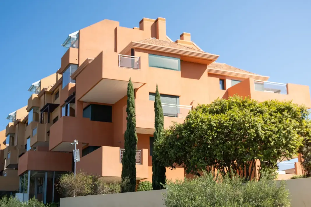 Un edificio de apartamentos moderno de estilo mediterráneo con paredes de terracota y vegetación exuberante bajo un cielo claro.